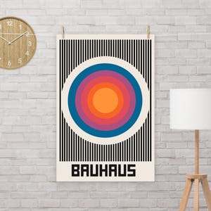 Puede incluir: Un póster con un fondo de rayas blancas y negras y un gran círculo colorido en el centro. El círculo tiene anillos concéntricos de naranja, rojo, rosa, morado y azul. La palabra "BAUHAUS" está impresa en negro en la parte inferior del póster.