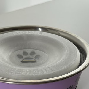 May include: A stainless steel pet bowl with a lavender-colored base and a paw print design. The bowl has a gray insert with a paw print in the center. The rim of the bowl is silver.