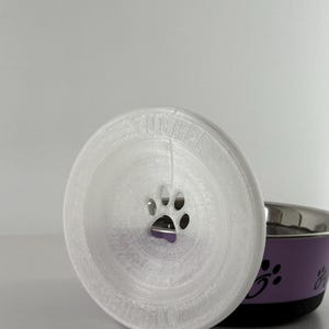 May include: A white, 3D-printed pet food bowl lid with a paw print cutout. The lid is circular and sits in front of a purple and silver pet bowl with paw print designs.