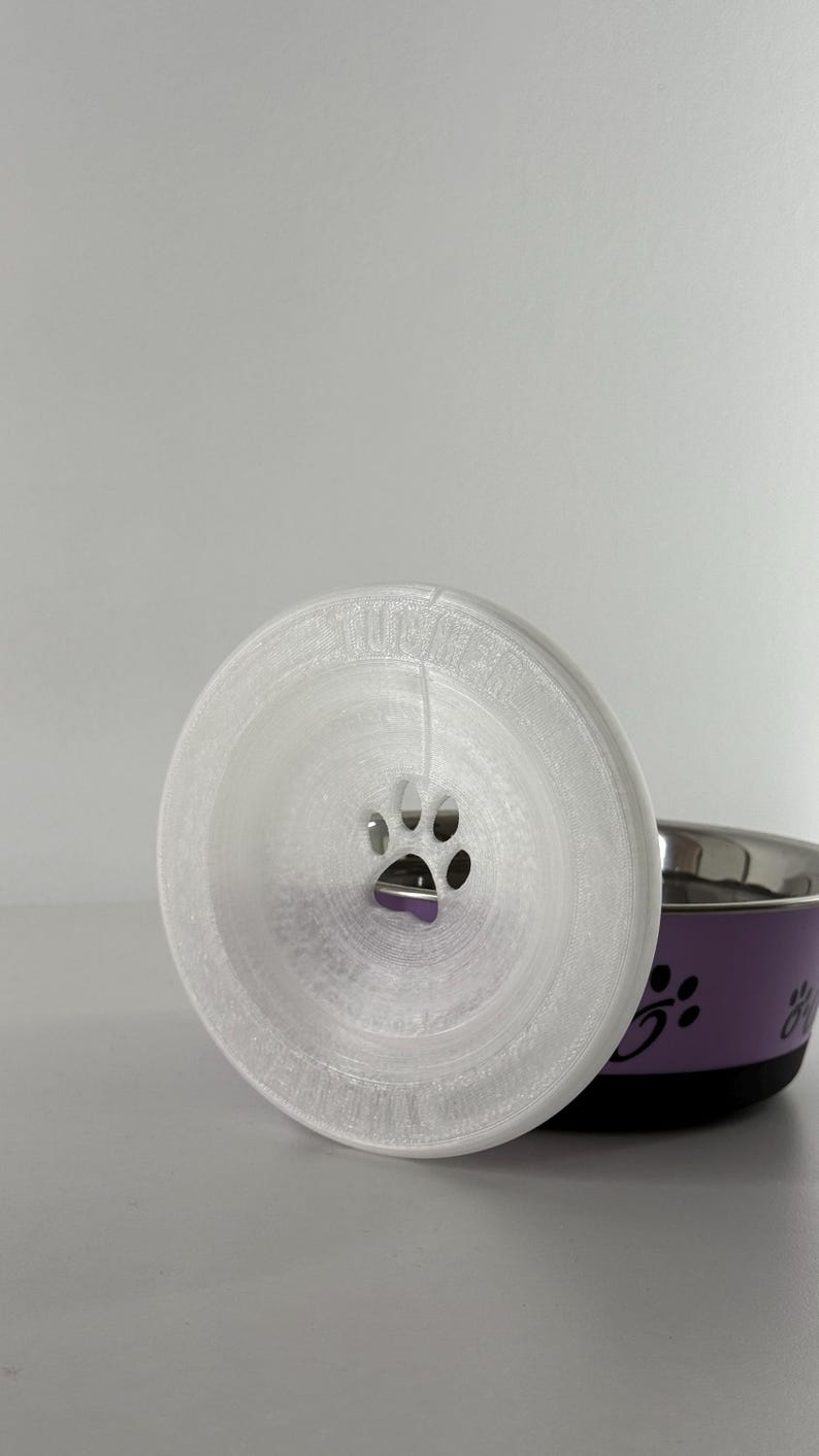 May include: A white, 3D-printed pet food bowl lid with a paw print cutout. The lid is circular and sits in front of a purple and silver pet bowl with paw print designs.
