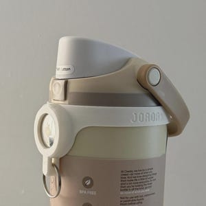 Könnte beinhalten: Eine beige-weiße Wasserflasche mit Klappdeckel und Griff. Auf der Flasche steht das Wort "JORDAN". Die Flasche hat auch ein Etikett mit der Aufschrift "BPA-FREI", "LEICHT ZU REINIGEN" und "BECHERHALTER-FREUNDLICH".