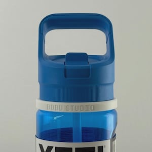 Puede incluir: Botella de agua YETI Yonder azul con asa y tapa azules. La botella tiene una banda blanca con "BOBU STUDIO" impresa. La botella está etiquetada como "YETI YONDER 600 ml".