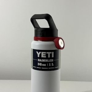 May include: White YETI Rambler water bottle with a black handle and red accents. The bottle has a 36 oz capacity, with the YETI logo and product details printed on the front. The bottle is designed for outdoor use.