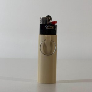 May include: A beige lighter holder with a silver ring attached. A black BIC lighter with a red button is inside the holder. The holder is cylindrical and made of a smooth material.