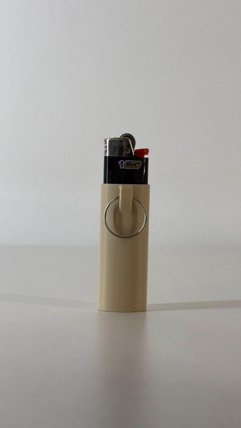 May include: A beige lighter holder with a silver ring attached. A black BIC lighter with a red button is inside the holder. The holder is cylindrical and made of a smooth material.