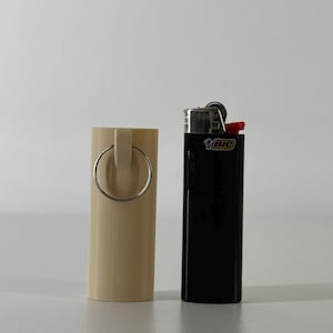 May include: A beige cylindrical lighter holder with a silver ring handle stands next to a black BIC lighter with a red top. The items are on a white surface.