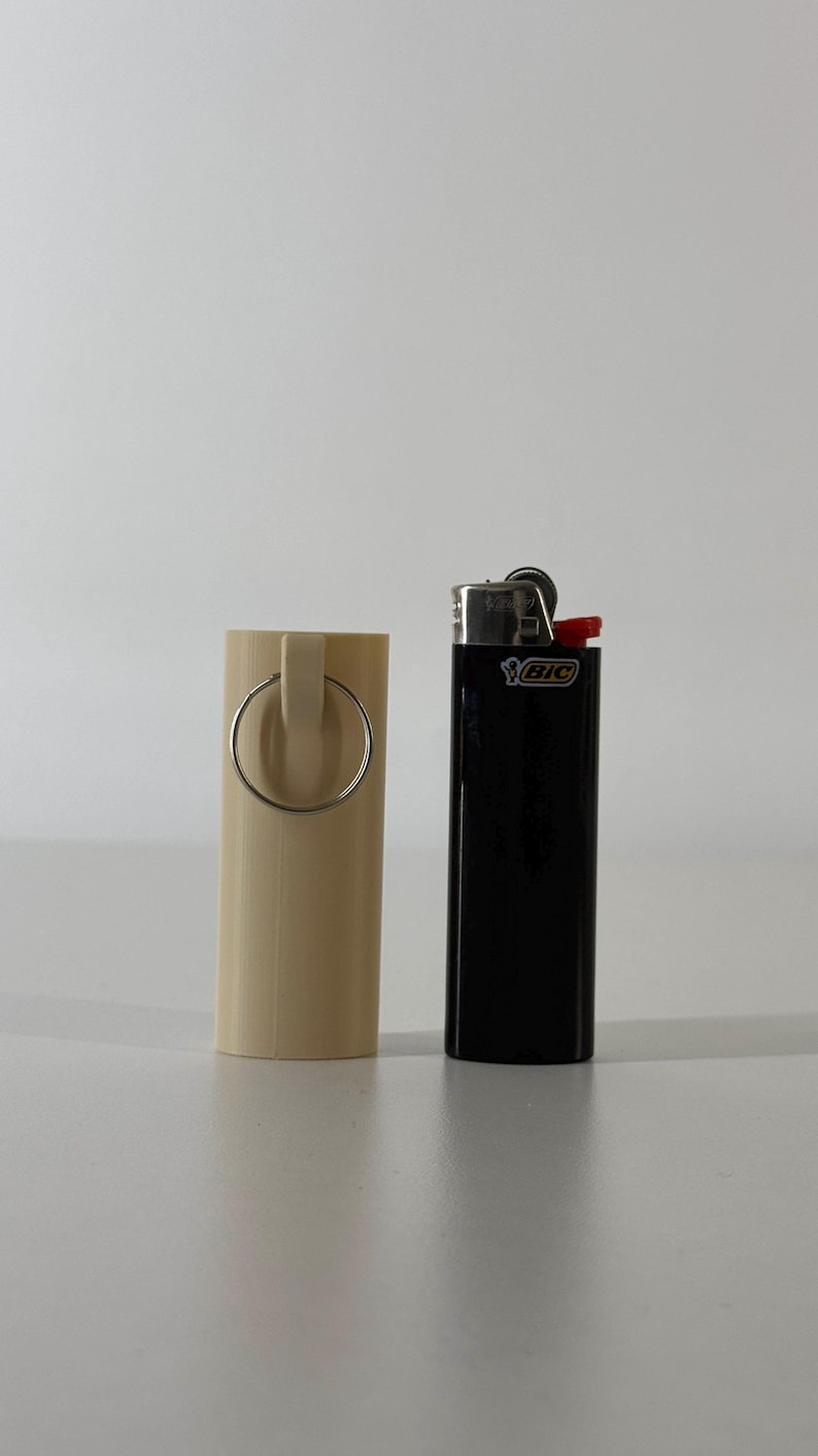 May include: A beige cylindrical lighter holder with a silver ring handle stands next to a black BIC lighter with a red top. The items are on a white surface.