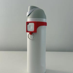 Puede incluir: Una botella de agua Owala blanca con tapa gris y accesorio Tile rojo. La botella tiene un asa y un accesorio rojo con un anillo plateado. La palabra "owala" está impresa en la botella.