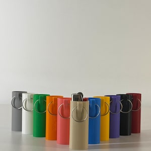 May include: A collection of colorful lighter cases in various shades, including gray, white, green, orange, coral, beige, blue, yellow, purple, black, and burgundy. Each case has a silver ring attached.