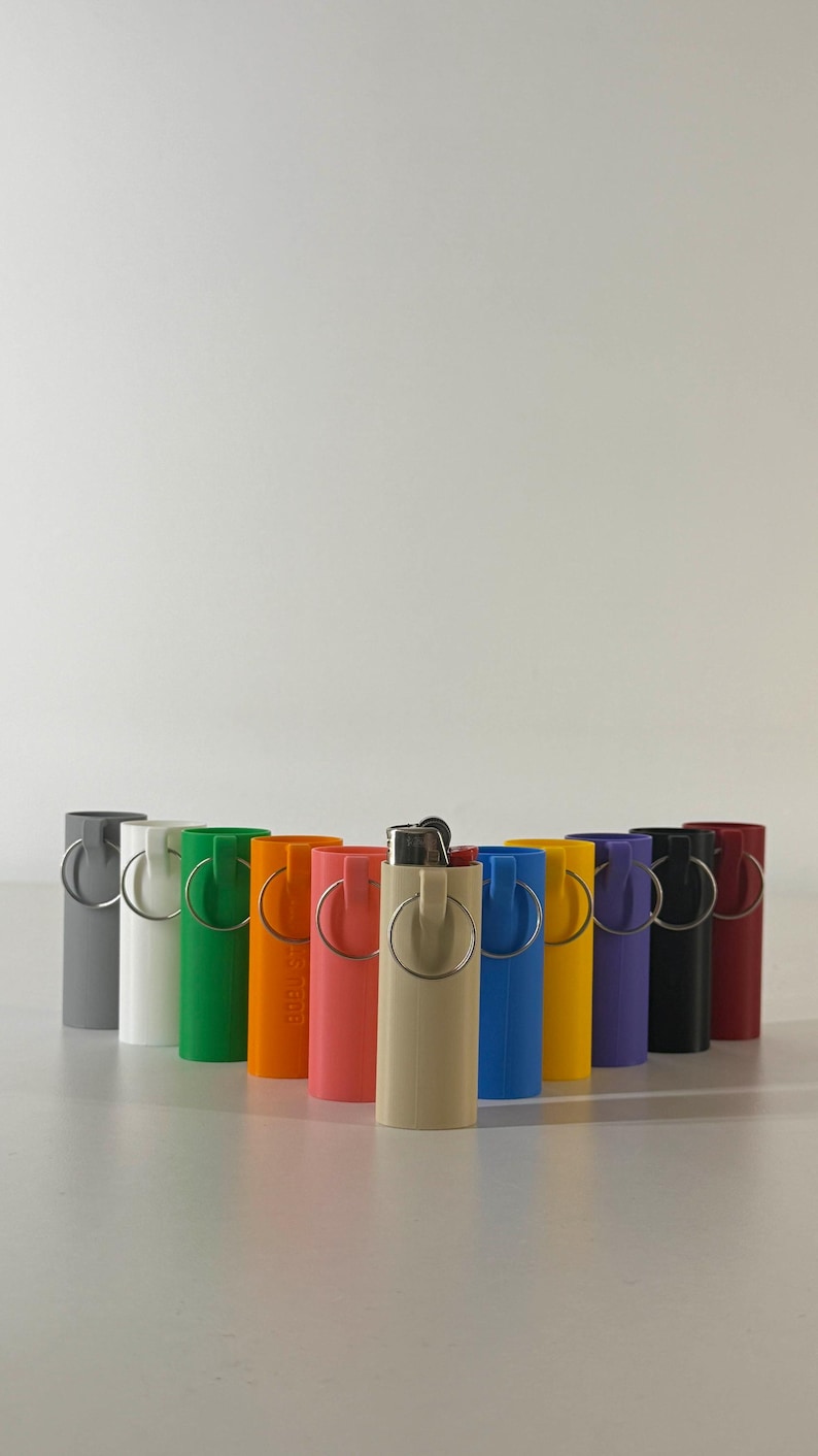 May include: A collection of colorful lighter cases in various shades, including gray, white, green, orange, coral, beige, blue, yellow, purple, black, and burgundy. Each case has a silver ring attached.