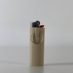 May include: A beige lighter case with a silver ring attached. The top of the lighter is visible, with a red ignition button. The case is cylindrical and appears to be made of plastic.