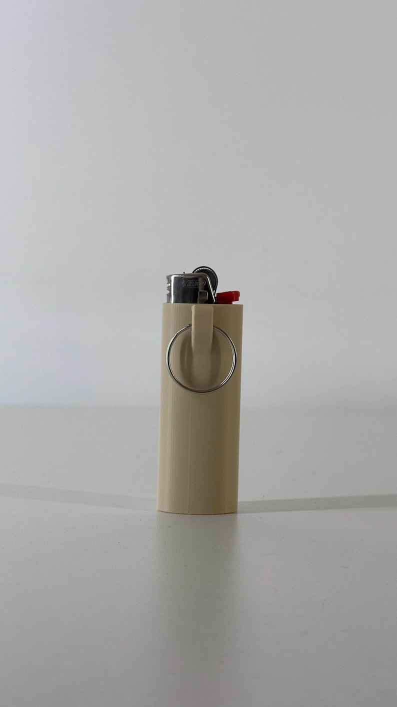 May include: A beige lighter case with a silver ring attached. The top of the lighter is visible, with a red ignition button. The case is cylindrical and appears to be made of plastic.