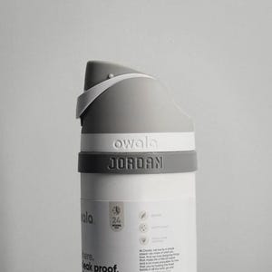 May include: A white and grey Owala water bottle with the name "JORDAN" printed on the side. The bottle has a grey handle and a white lid. The bottle is leak proof, insulated, and has a built-in straw.