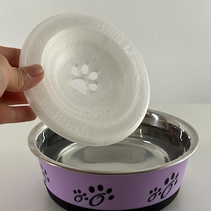 May include: A stainless steel pet bowl with a lavender band featuring black paw prints. A clear, circular lid with a paw print design is held above the bowl. The bowl has a black base.