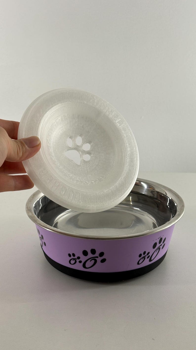 May include: A stainless steel pet bowl with a lavender band featuring black paw prints. A clear, circular lid with a paw print design is held above the bowl. The bowl has a black base.