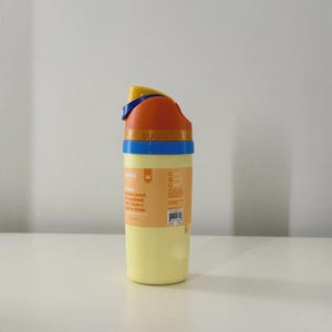 Puede incluir: Una botella de agua amarilla con una tapa azul, naranja y amarilla. La botella tiene las palabras "owala" y "BUBU" impresas. La botella tiene una capacidad de 710 ml y una pajita incorporada.