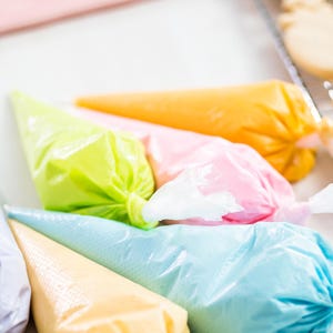 Op de afbeelding: Vijf spuitzakken gevuld met verschillende gekleurde frosting. De frostingkleuren zijn paars, geel, blauw, roze en oranje. De zakken zijn in een halve cirkel op een wit oppervlak gerangschikt.