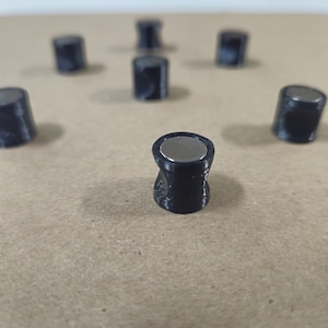 May include: A collection of small, black cylindrical objects with a silver disc on top. The objects are arranged on a light brown surface. The objects appear to be magnets.