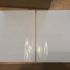 Può includere: Due fogli rettangolari di carta bianca, possibilmente materiale da imballaggio, sono appoggiati uno accanto all'altro su una superficie di legno. Un cartone è visibile sullo sfondo. La carta ha una qualità leggermente traslucida.