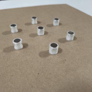 May include: Seven white cylindrical magnets with black centers are arranged in a circular pattern on a brown surface.
