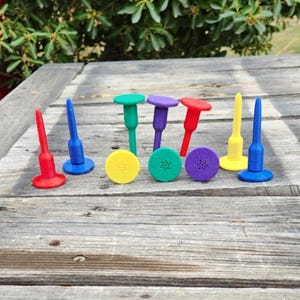 May include: A selection of vibrant golf tees and ball markers in red, blue, green, purple, and yellow. The tees have a peg design, while the markers are round with a textured surface. The items are arranged on a weathered wooden surface.