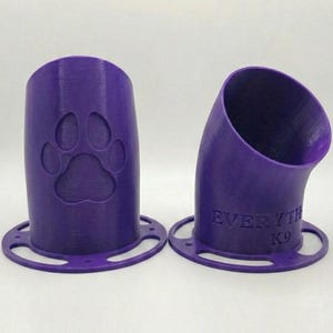 Op de afbeelding: Twee paarse, 3D-geprinte hondenkoekjesdispensers. Eén heeft een pootafdruk, de andere de tekst "EVERYTH K9". Beide hebben een cirkelvormige basis met gaten. De dispensers zijn schuin en cilindrisch.
