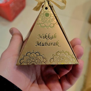 Peut inclure: Bo&icirc;te cadeau dor&eacute;e en forme de pyramide avec des motifs floraux d&eacute;coratifs et l'inscription "Nikkah Mubarak". La bo&icirc;te est attach&eacute;e avec un ruban dor&eacute;. La bo&icirc;te est tenue dans la main d'une personne.