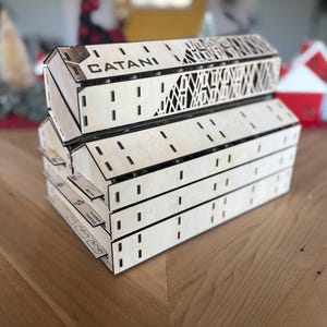 May include: Stacked wooden game boxes with the word "CATANI" etched on the top. The boxes are light-colored with decorative cutouts and slots. The game boxes are on a wooden surface.