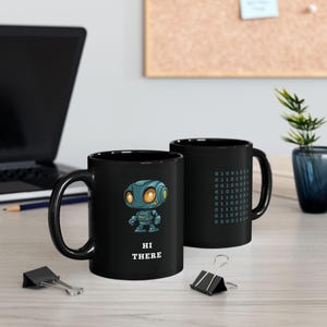 Puede incluir: Dos tazas de cerámica negra con un gráfico de robot y el texto "Hi There". Una taza tiene un patrón de código binario en el lateral.