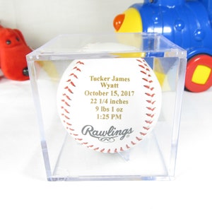 May include: A white baseball with gold lettering in a clear plastic display case. The baseball is inscribed with "Tucker James Wyatt", "October 15, 2017", "22 1/4 inches", "9 lbs 1 oz", and "1:25 PM". The baseball is a Rawlings brand.