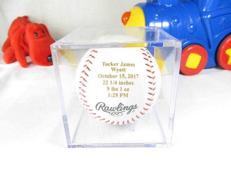 May include: A white baseball with gold lettering in a clear plastic display case. The baseball is inscribed with "Tucker James Wyatt", "October 15, 2017", "22 1/4 inches", "9 lbs 1 oz", and "1:25 PM". The baseball is a Rawlings brand.