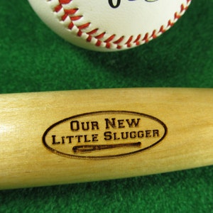May include: A wooden baseball bat with the words "Our New Little Slugger" engraved on it.