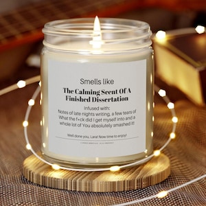 May include: A lit glass jar candle on a wooden coaster, surrounded by string lights. The candle label reads "Smells like The Calming Scent Of A Finished Dissertation." The text continues with "Infused with: Notes of late nights writing..."