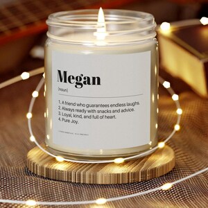 May include: A lit candle in a clear glass jar with a label that reads "Megan" and a definition. The candle sits on a wooden coaster, surrounded by string lights. The background is blurred.