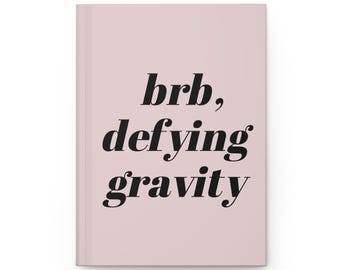 May include: A hardcover journal with a pale pink cover. The words "brb, defying gravity" are printed in black, bold font across the front. The journal is closed and appears new.