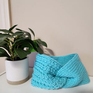 May include: A turquoise knitted headband with a twisted knot design. The headband is made of a chunky knit material and is displayed on a white surface. A potted plant is visible in the background. The text "ThreeThreads" is at the bottom left.