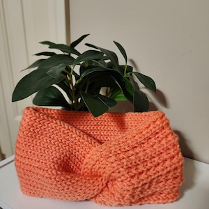 May include: A coral-colored, knitted headband with a twisted knot design. The headband is shown with a green plant inside. The text "ThreeThreads" is at the bottom left.