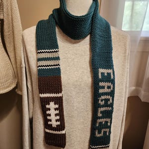 May include: A teal, brown, white, and grey knitted scarf featuring a football graphic on one end and the word EAGLES knitted in white on the other. The scarf has horizontal stripes in grey, white, and black near the ends and a textured knit pattern.