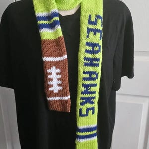 Puede incluir: Una bufanda de punto verde lima con un gráfico de balón de fútbol americano marrón con cordones blancos en un extremo y la palabra SEAHAWKS en letras azules en el otro. La bufanda incluye rayas horizontales azules y blancas en los extremos.