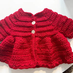 Op de afbeelding: Een rood gebreid babyvest met knoopsluiting. Het vest heeft een gerimpelde zoom en een gegolfde halslijn.