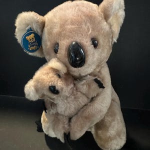 Peut inclure: Un ours en peluche koala brun tenant un plus petit ours en peluche koala brun. Le plus grand koala a un nez noir et des yeux noirs. Le plus petit koala a un nez noir et des yeux noirs. Le plus grand koala a une étiquette bleue sur son oreille qui dit "Carry Me".