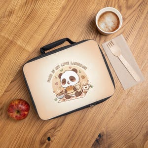 Könnte beinhalten: Eine rechteckige Lunchtasche mit schwarzem Griff und Reißverschluss, die einen Cartoon-Panda beim Essen zeigt. Der Text "FOOD IS MY LOVE LANGUAGE" ist auf die Tasche gedruckt. Ein Apfel, eine Kaffeetasse und eine Gabel sind ebenfalls zu sehen.