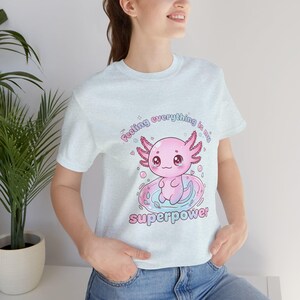 Puede incluir: Camiseta azul claro con un diseño de ajolote de dibujos animados. El ajolote rosa tiene ojos grandes y está sentado en un anillo rosa y azul. El texto de la camiseta dice: "Feeling everything is my superpower."