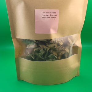 Peut inclure: Un sac en papier brun avec une fenêtre transparente, rempli d'herbes séchées. Une étiquette rose sur le sac indique "fey zanmorèt (turkey berry) baya de pavo."