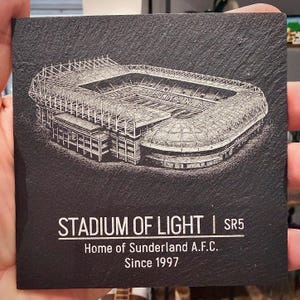 Puede incluir: Un posavasos cuadrado de pizarra negra con una detallada ilustraci&oacute;n grabada con l&aacute;ser en blanco del Stadium of Light. El texto dice STADIUM OF LIGHT | SR5, Home of Sunderland A.F.C., Since 1997. La superficie tiene una textura natural y rugosa.