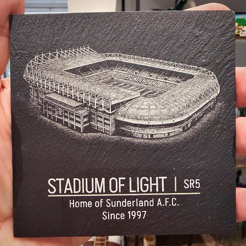 Puede incluir: Un posavasos cuadrado de pizarra negra con una detallada ilustraci&oacute;n grabada con l&aacute;ser en blanco del Stadium of Light. El texto dice STADIUM OF LIGHT | SR5, Home of Sunderland A.F.C., Since 1997. La superficie tiene una textura natural y rugosa.