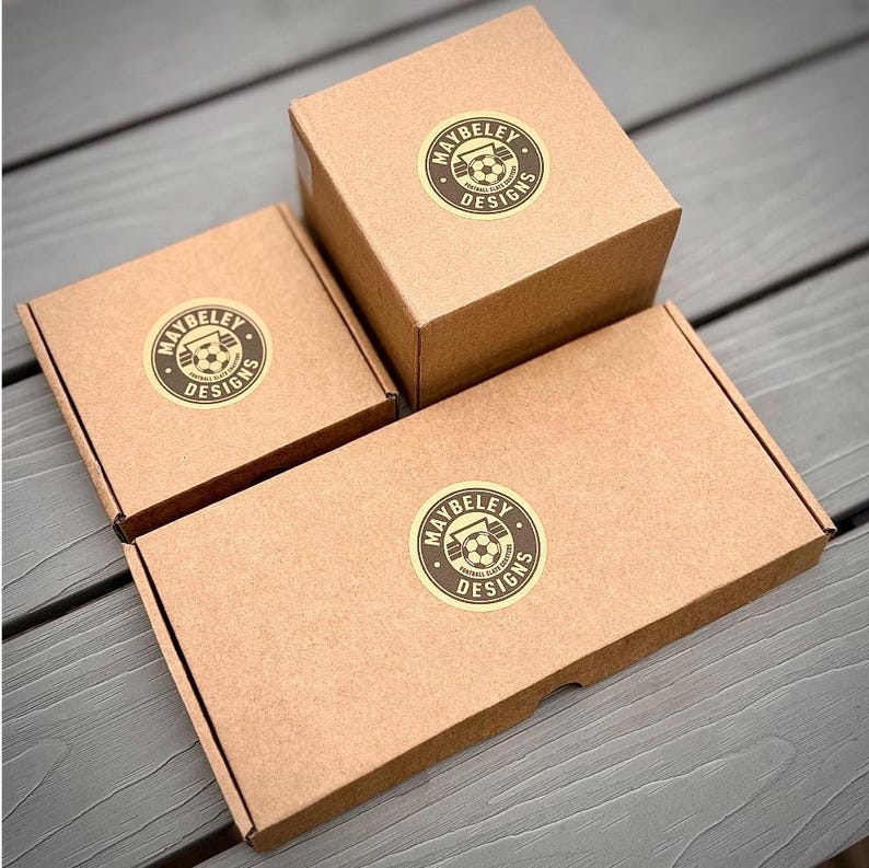 Puede incluir: Tres cajas de cart&oacute;n marr&oacute;n de diferentes tama&ntilde;os, cada una con una pegatina circular con el texto MAYBELEY DESIGNS y un gr&aacute;fico de un bal&oacute;n de f&uacute;tbol. Las cajas est&aacute;n dispuestas en un grupo, mostrando una caja cuadrada y una rectangular.