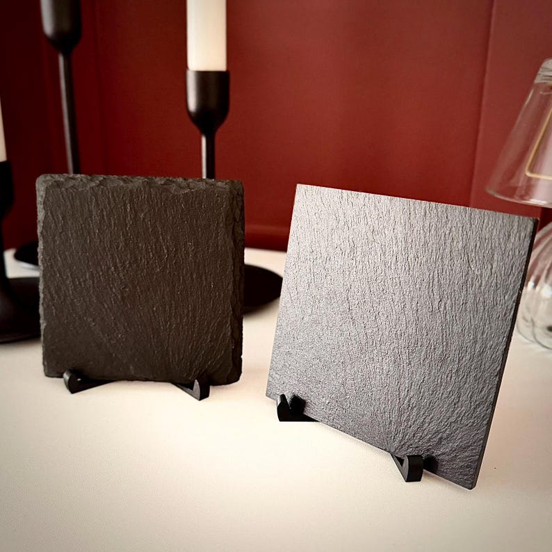 Puede incluir: Dos posavasos cuadrados de pizarra negra con bordes rugosos y texturizados, cada uno expuesto en posici&oacute;n vertical sobre un peque&ntilde;o soporte de pl&aacute;stico negro. Las superficies de piedra oscura presentan un grano natural e irregular.