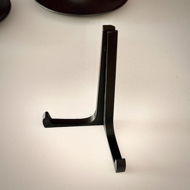 Puede incluir: Un soporte de exhibici&oacute;n negro en forma de L que cuenta con un respaldo vertical y una base horizontal con un peque&ntilde;o gancho curvado hacia arriba en el borde frontal para sostener objetos en posici&oacute;n vertical. Acabado mate y liso.
