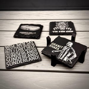 Puede incluir: Cuatro posavasos cuadrados de pizarra negra con bordes irregulares. Muestran el Stadium of Light, una pantera con 'TIL THE END', letras sobre Sunderland y un retrato de Granit Xhaka, en un soporte negro sobre una superficie de madera.
